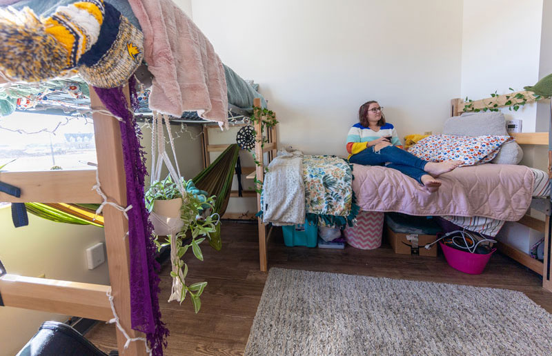 Female student sitting on a bed in a dorm room in Gold Hall. There are two beds and the room is decorated.
